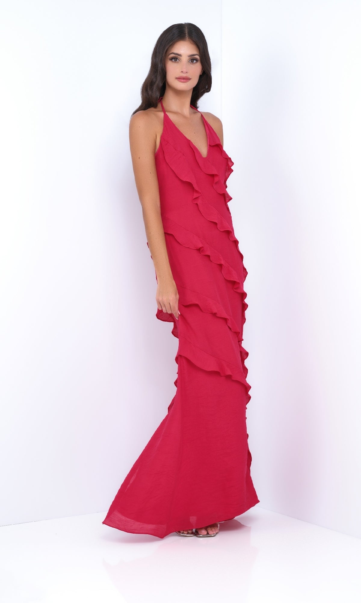 A woman stands against a white backdrop wearing the Remi by Velvi—a V-Neck Halter Ruffled Long Prom Dress in striking red, featuring elegant sleeveless details.
